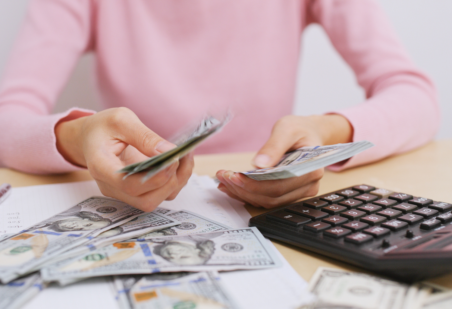 Fashionable dressed women in a pink sweater counting money and calculating her capital expenditure on a brown wood desk.